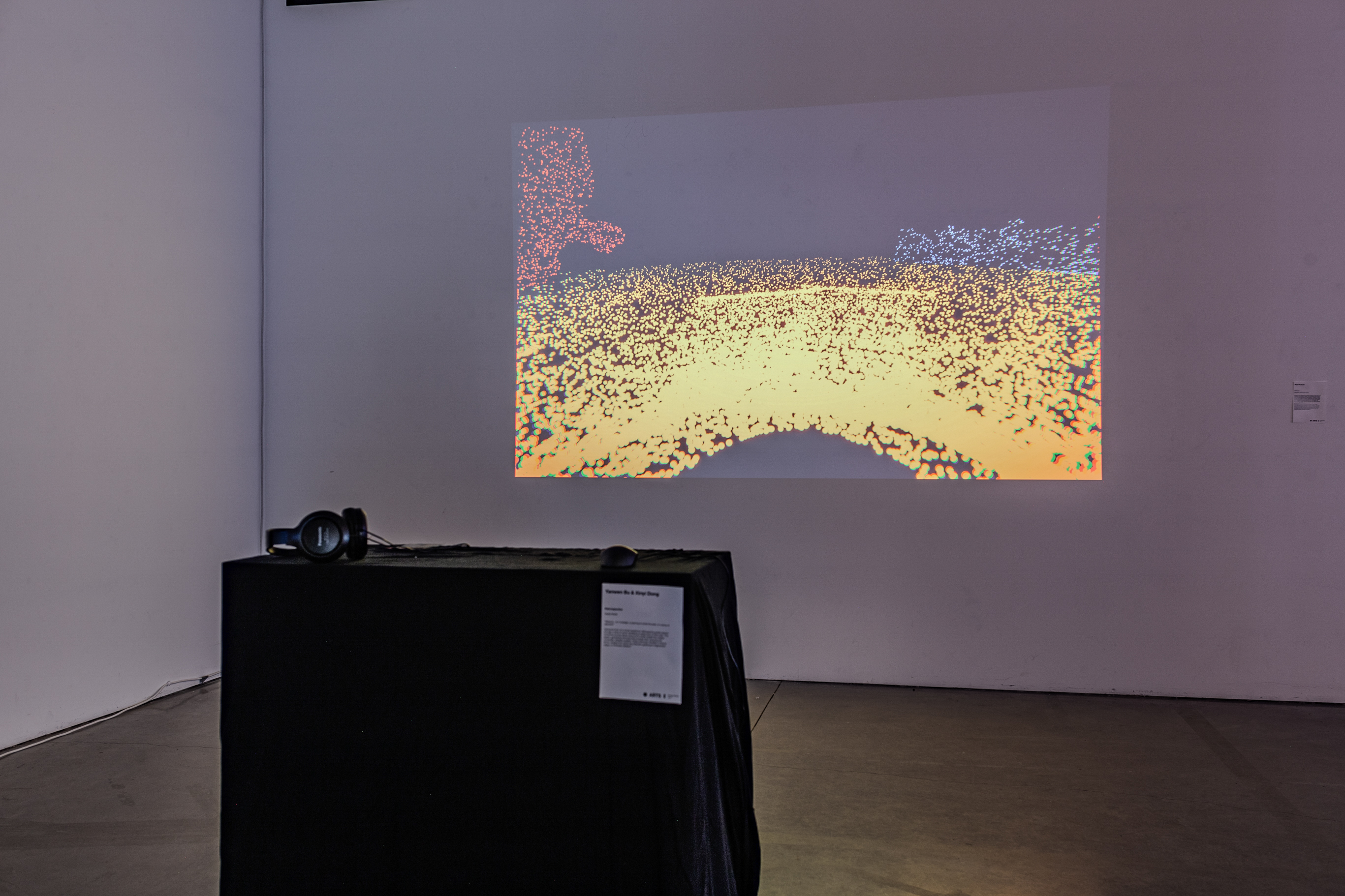 A projection of a virtual interactive game onto a white wall. In the foreground is a black tabl with headphones, keyboard, a mouse.